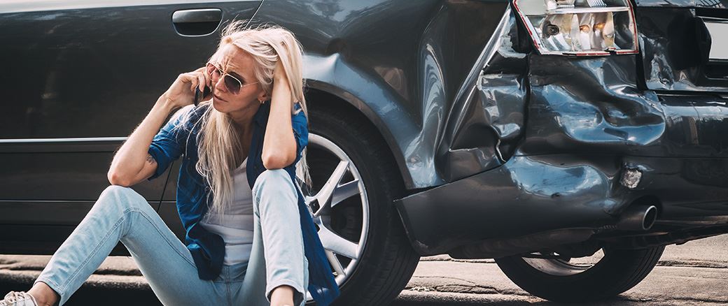 Woman on her phone sitting on the ground next to her damaged car.